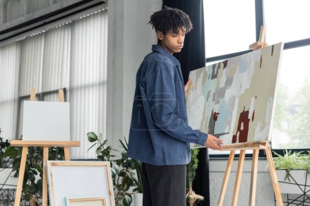 Young african american artist taking painting from easel in workshop 