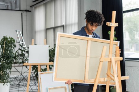 Young african american artist holding canvas near wooden easel in studio 