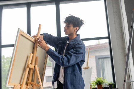 Téléchargez les photos : Low angle view of young african american artist adjusting canvas on wooden easel - en image libre de droit