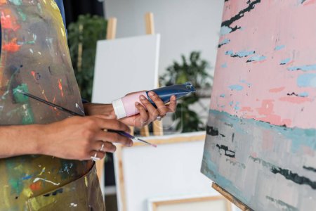 Vue recadrée de l'artiste afro-américain en tablier tenant le tube de peinture et le pinceau près du dessin 
