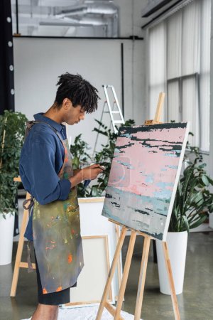 Téléchargez les photos : Side view of african american artist in apron holding paintbrush and paint near drawing in studio - en image libre de droit