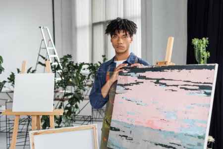 Junger afrikanisch-amerikanischer Künstler in Schürze blickt in Werkstatt in die Kamera 