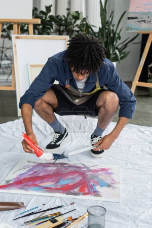 African american artist pouring paint on canvas near paintbrushes on cloth in studio
