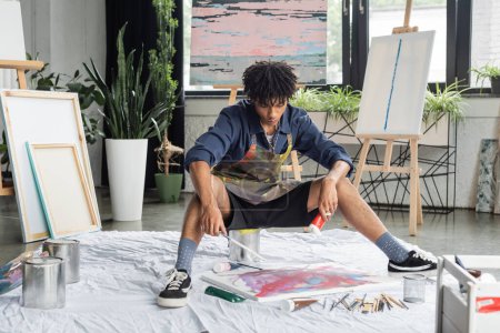 Junge afrikanisch-amerikanische Künstlerin hält Farbe und Pinsel in der Nähe von Leinwand auf dem Boden in der Werkstatt 