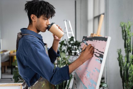 Junge afrikanisch-amerikanische Künstlerin trinkt Coffee to go und malt in Werkstatt auf Leinwand 