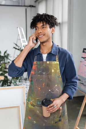 Smiling african american artist in dirty apron talking on smartphone and holding coffee to go in studio 