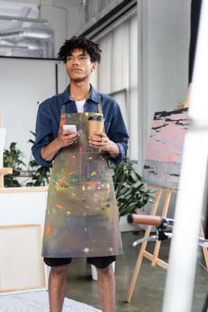 Pensive african american artist in apron holding smartphone and coffee in workshop 