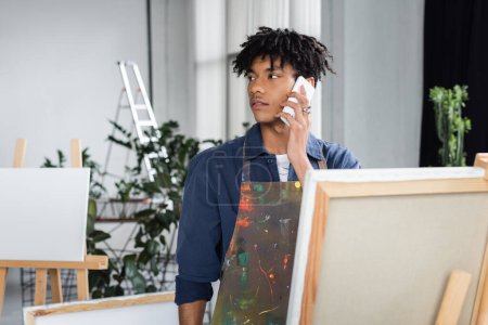 Joven artista afroamericano en delantal hablando por celular en taller 