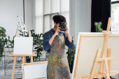 Young african american artist in apron taking photo on vintage camera near canvas in studio  Stickers #638029670