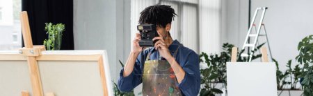 Artiste afro-américain prenant des photos sur caméra vintage près de la toile sur chevalet en studio, bannière 