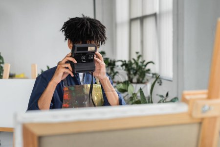 Artista afroamericano tomando fotos en cámara vintage cerca de lienzo borroso en estudio 
