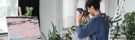 Joven artista afroamericano tomando fotos en cámara retro cerca del dibujo en el taller, pancarta 
