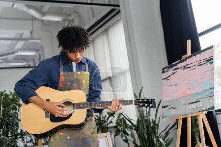 Joven artista afroamericano en delantal tocando guitarra acústica en taller 