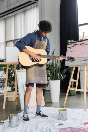 African american artist playing acoustic guitar near paints and easels in workshop 