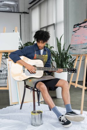 Artista afroamericano en delantal tocando guitarra acústica cerca del dibujo y lienzos en estudio 