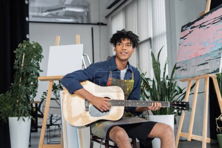 Sonriente artista afroamericano en delantal sucio tocando guitarra acústica cerca de caballetes en estudio 