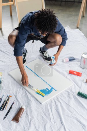 African american artist in apron painting with spatula on canvas in studio 
