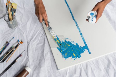 Téléchargez les photos : Cropped view of african american artist painting with spatula near paintbrushes in studio - en image libre de droit