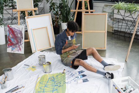 Artista afroamericano usando smartphone cerca de pinturas en el piso en el estudio 