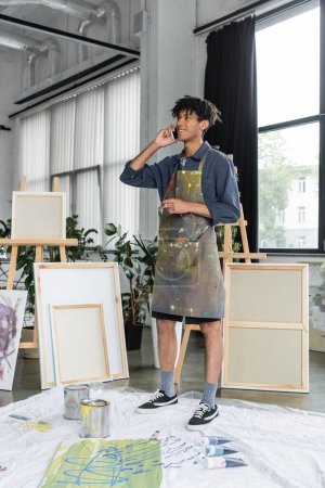 Smiling african american artist in apron talking on smartphone near paints in studio 