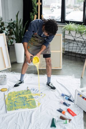 Foto de Artista afroamericano vertiendo pintura sobre tela cerca de lienzo en estudio - Imagen libre de derechos