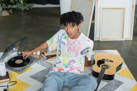 Téléchargez les photos : Jeune artiste afro-américain utilisant un tourne-disque près de la guitare acoustique et des toiles en studio - en image libre de droit