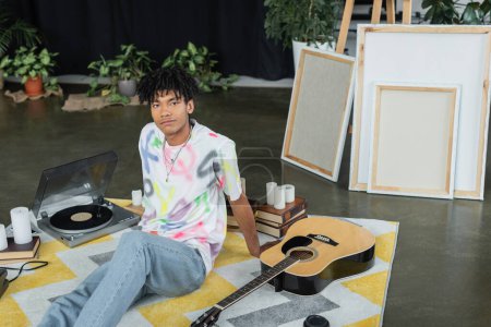 Foto de Artista afroamericano mirando cámara cerca de guitarra acústica y lienzos en taller - Imagen libre de derechos