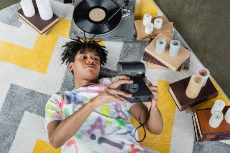 Téléchargez les photos : Vue du dessus de l'artiste afro-américain tenant une caméra vintage près des livres et un tourne-disque en studio - en image libre de droit