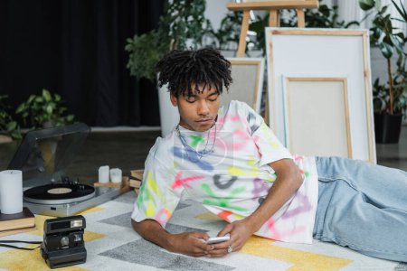 Téléchargez les photos : Artiste afro-américain utilisant un smartphone près de la caméra vintage et des toiles floues - en image libre de droit