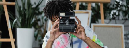 Joven artista afroamericano tomando fotos en cámara vintage en el estudio, pancarta 