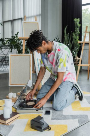 African american artist using record player near books and canvases in workshop 