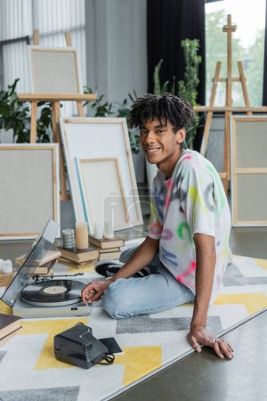 Lächelnder afrikanisch-amerikanischer Künstler blickt in Werkstatt in die Kamera neben Plattenspieler und Büchern 