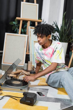 Joven artista afroamericano usando tocadiscos cerca de libros y cámara vintage en taller 