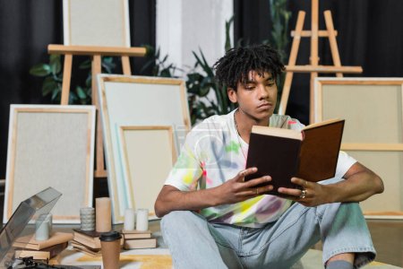 African american artist reading book near record player and canvases in workshop 