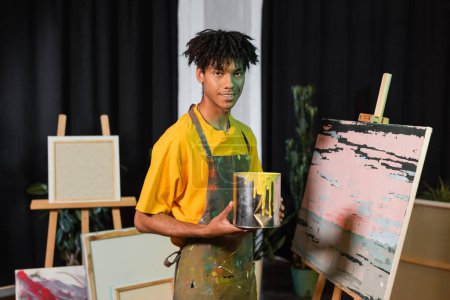 Sonriente artista afroamericano sosteniendo tarro con pintura cerca de dibujo en taller 