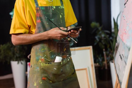Cropped view of african american artist holding paintbrush and using smartphone in studio 