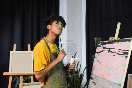 Artiste afro-américain pensif tenant peinture et pinceau près du dessin en atelier 