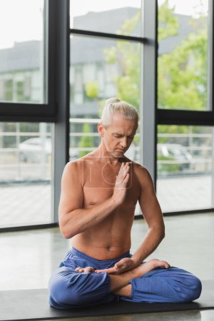 man with closed eyes sitting in lotus pose on yoga mat in studio 