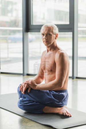 grey haired man in pants sitting in twisting yoga pose on mat and looking at camera