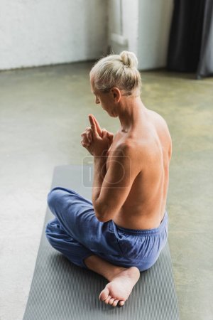 shirtless man in pants sitting in twisting yoga pose and doing crown chakra mudra