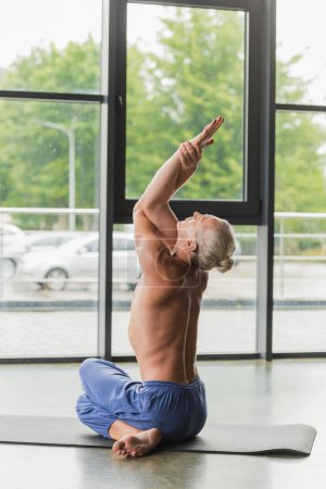 shirtless man in blue pants sitting in twisting yoga pose and raising arms 