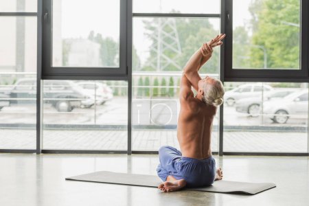 shirtless man in blue pants sitting in twisting yoga pose and raising arms while stretching back 