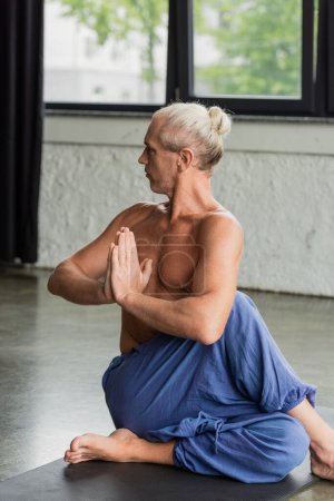 Photo for Grey haired man in pants sitting in half lord of fishes pose and doing anjali mudra - Royalty Free Image