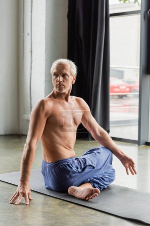 shirtless and grey haired man in pants sitting in half lord of fishes pose in yoga studio 