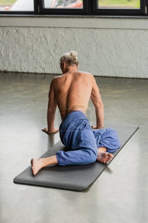 hombre descalzo y de pelo gris en pantalones sentados con las piernas retorcidas mientras que estira detrás en la estera del yoga 