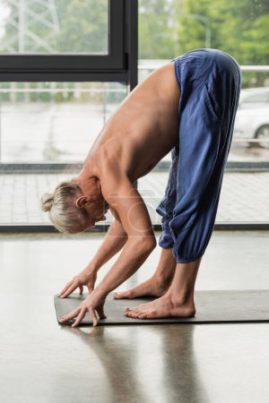 shirtless and grey haired man doing standing forward bend pose on yoga mat 