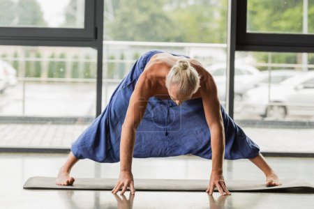 grey haired man doing wide legged forward bend on yoga mat in studio 