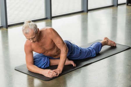 Téléchargez les photos : Homme aux cheveux gris faisant demi pose de pigeon sur le tapis dans un studio de yoga - en image libre de droit