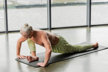 grey haired man doing seated forward bend on yoga mat in studio 