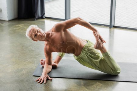 shirtless man doing supine spinal twist yoga pose in studio 